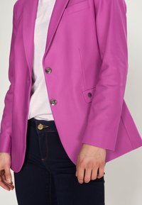 Fuchsiový blejzr s propracovaným designem, dvěma kovovými knoflíky a jednou přední kapsou, ve spojení s tmavými džíny a světlou košilí.