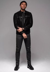 Chaqueta de cuero negra con bolsillos con cremallera, combinada con pantalones slim-fit negros y botas de cordones. El modelo se encuentra de pie frente a un fondo gris.