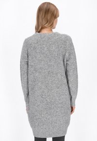 O femeie cu părul lung, purtând o rochie-swetere gri oversized dintr-un material pufos, arătată din spate pe un fundal alb simplu.