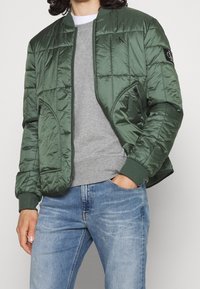 Zielona pikowana kurtka typu bomber z zamkiem z przodu, bocznymi kieszeniami, ściągaczami na mankietach i naszywką z logo na rękawie. Nosi się ją na szarej bluzie.