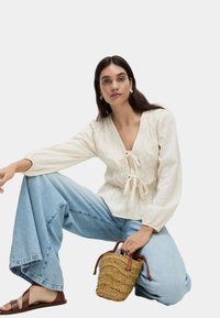 Blusa crema con dettagli a fiocco e motivo testurizzato, abbinata a jeans in denim azzurro a gamba larga. Accessoriata con una borsa in rafia.