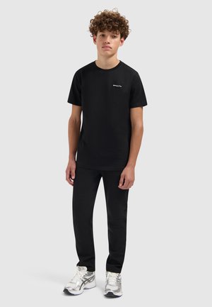 Zwarte katoenen T-shirt met een ronde hals en een klein wit logo op de borst, gecombineerd met zwarte broek en zilveren sport schoenen.
