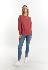 Červená mikina s dlhými raglánovými rukávmi, rebrovanými manžetami a lemom, v kombinácii s modrými skinny džínsami a bielymi teniskami na svetlom pozadí.