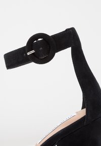 Chaussure à talon haut en daim noir avec une bride à la cheville et une boucle circulaire. Présente une doublure intérieure lisse et un design profilé élégant.