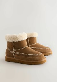 Bruna mockakängor med en fluffig krämfärgad pälskrage, dekorativ sömnad och en strukturerad gummisula, med en rundad tådesign.