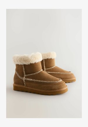 Stivali ankle in suede marrone con un morbido colletto in pelliccia crema, cuciture decorative e una suola in gomma testurizzata, caratterizzati da una punta arrotondata.