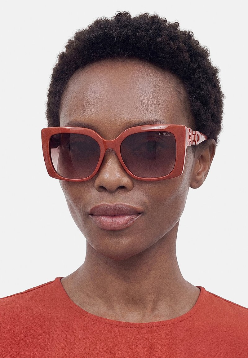 Personne aux cheveux courts et bouclés portant de grandes lunettes de soleil rouges carrées et un haut rouge assorti sur un fond neutre.