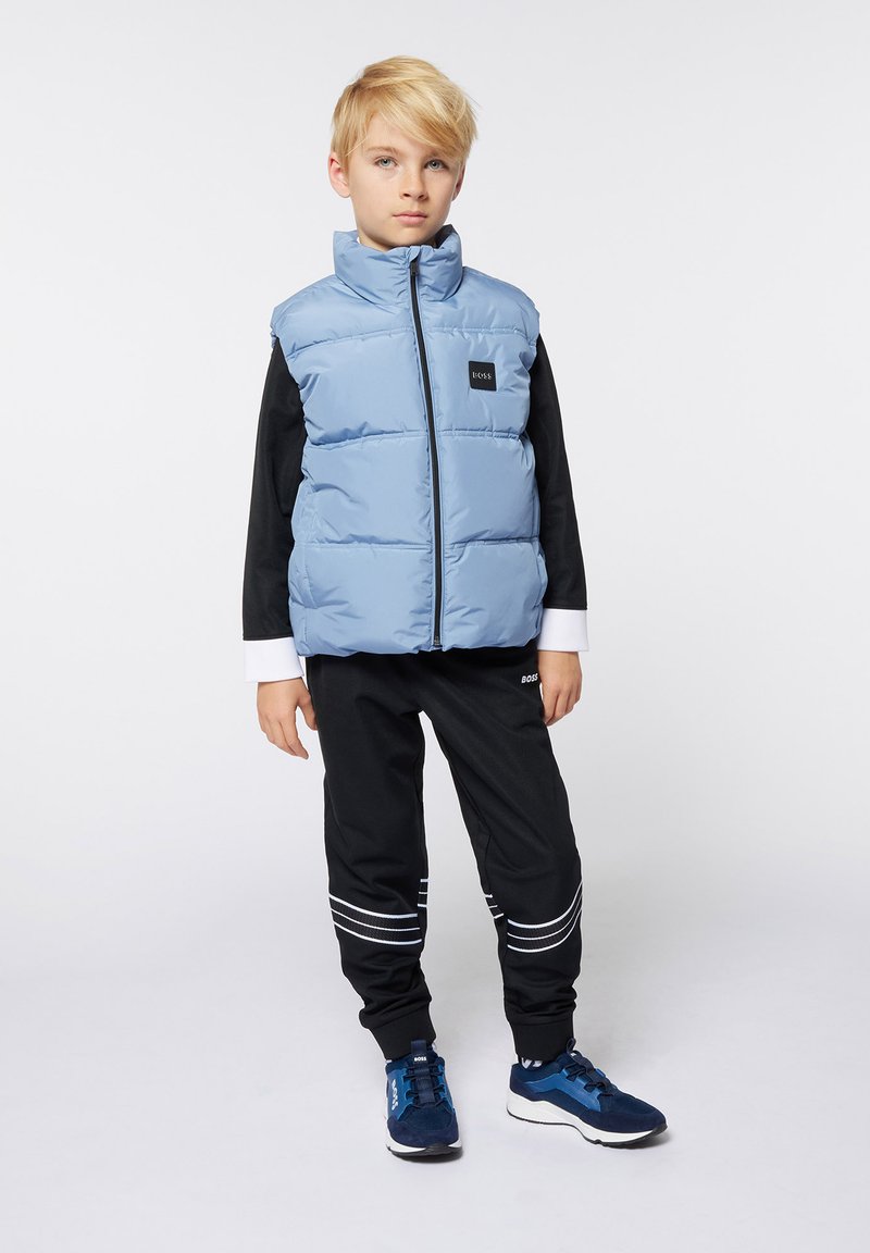 Lichtblauwe puffervest met een hoge kraag en een rits aan de voorkant, gelaagd over een zwart langemouwen shirt, gecombineerd met zwarte broek en blauwe sneakers.