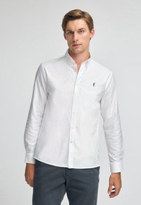 Camicia bianca a maniche lunghe con colletto button-down, caratterizzata da una piccola figura nera ricamata sul lato sinistro del petto e una texture liscia.