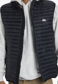 Padded black vest with horizontal quilting, front zipper, and logo patch on the chest. Worn over a gray hoodie, featuring a soft texture.