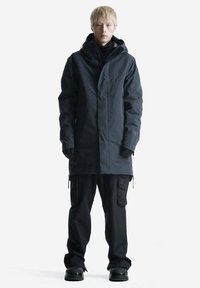 Parka nera impermeabile con cappuccio, dotata di zip frontale, tasche laterali e tessuto testurizzato. Abbinata a pantaloni cargo neri larghi e stivali.