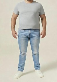 Homme portant un t-shirt gris clair, un jean bleu clair et des baskets blanches, se tenant devant un fond beige uni.