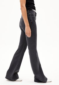 Zwart denim met uitlopende pijpen en een gladde textuur, met achterzakken en een hoge taille. Te zien in combinatie met witte schoenen.