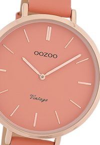 Montre ronde avec un boîtier en or rose, cadran rose mat, aiguilles fines et marqueurs d'heure minimalistes. Elle est dotée d'un bracelet en cuir rose clair.