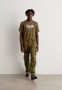 Olive green T-shirt with a bat graphic, paired with matching olive cargo pants. Black shoes with a red accent. Plain background.