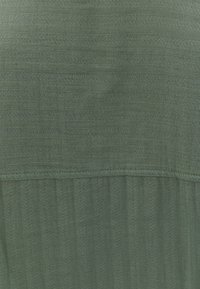 Tessuto verde texturizzato con strisce verticali e orizzontali, caratterizzato da una superficie liscia e una leggera lucentezza. Mostra una cucitura pulita all'orlo.