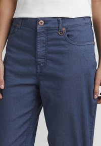 Los pantalones de mezclilla de un azul oscuro cuentan con un diseño de cinco bolsillos, botón de tono cobre y costuras sutiles a lo largo de las costuras. Tienen una textura suave y una forma ajustada.