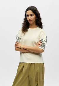 Femme aux cheveux bouclés et foncés portant un haut crème à manches courtes avec broderie florale noire et un pantalon plissé ample vert olive, bras croisés.