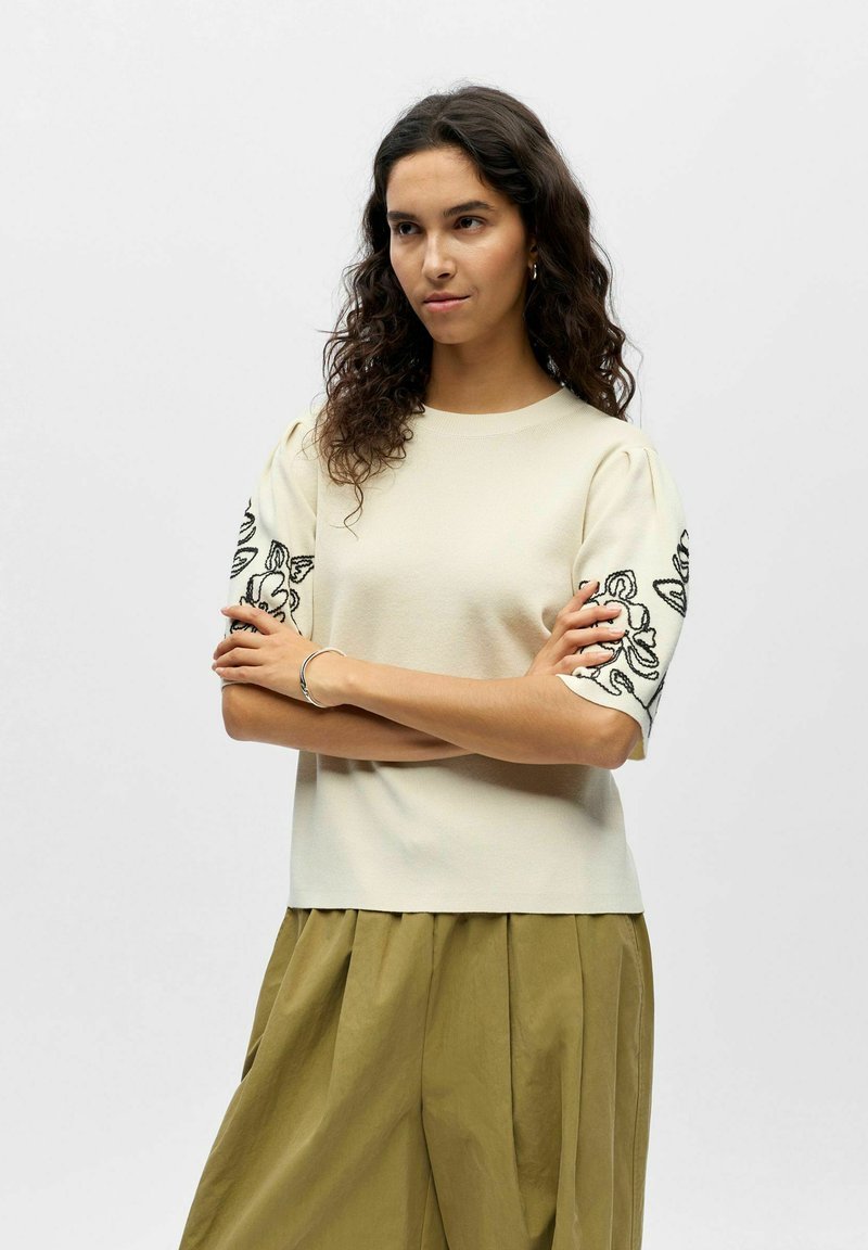 Femme aux cheveux bouclés et foncés portant un haut crème à manches courtes avec broderie florale noire et un pantalon plissé ample vert olive, bras croisés.