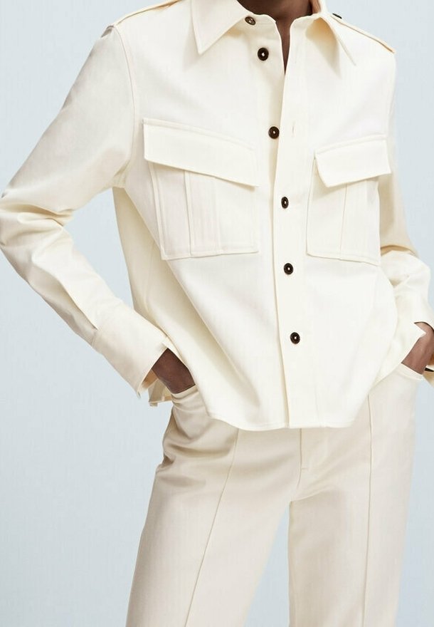 Chemise boutonnée couleur crème avec deux poches poitrine, boutons marron et coupe décontractée, portée avec un pantalon assorti couleur crème.