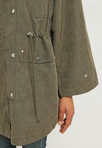 Zelená, lehká bunda s matnou texturou, vybavená tlakovýma knoflíkama, stahovací šňůrkou v pase a přední kapsou. Elegantní minimalistický design.