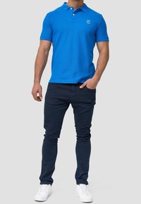 Polo bleu avec col et deux boutons, associé à un pantalon bleu marine et des baskets blanches, présenté sur un fond neutre.