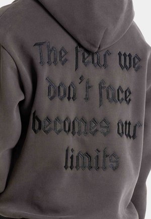 Sudadera con capucha gris que presenta un bordado negro texturizado con un texto gótico estilizado en la parte posterior, que dice: "El miedo que no enfrentamos se convierte en nuestros límites."