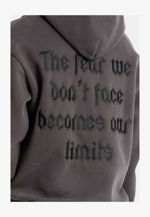 Sweat-shirt à capuche gris avec broderie noire texturée et texte gothique stylisé au dos, indiquant : "La peur que nous ne confrontons pas devient nos limites."