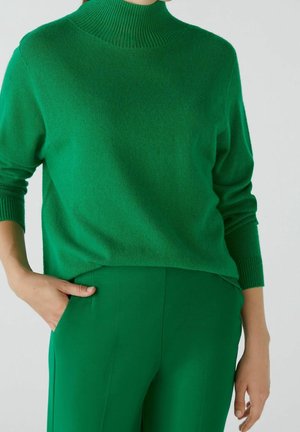 Femme portant un pull en maille vert à manches longues et un pantalon vert assorti, une main dans la poche, sur un fond uni.