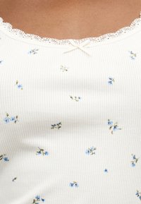 Tissu blanc côtelé avec petits motifs floraux bleus, bordure en dentelle le long de l'encolure, et un petit nœud décoratif au centre.