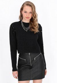 Schwarzer, verkürzter Pullover mit strukturiertem Muster und silbernen Hardware-Details an den Ärmeln. Kombiniert mit einem schwarzen Leder-Minirock mit Reißverschlüssen.
