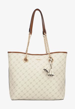 Sac fourre-tout crème avec petit motif géométrique, bordure marron, poignées en chaîne dorée et pendentif à pompon floral suspendu près du centre supérieur.