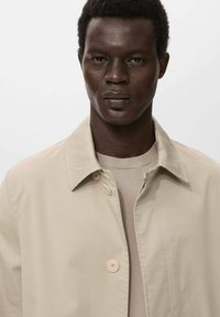 Beige leichte Jacke mit Kragen, mit zwei Knöpfen, über einem cremefarbenen Pullover getragen, vor einem neutralen Hintergrund präsentiert.