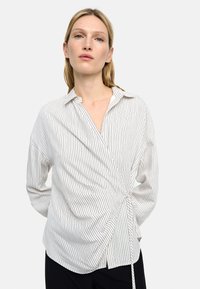 Koszula w paski wykonana z lekkiego materiału, z kopertowym fasonem i wiązaniem w pasie oraz klasycznym kołnierzykiem. Kolory obejmują biel i stonowane odcienie.