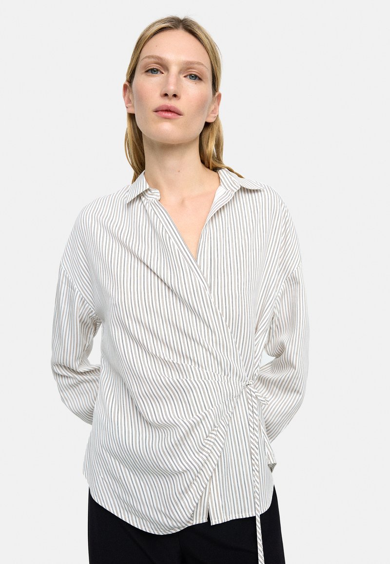 Koszula w paski wykonana z lekkiego materiału, z kopertowym fasonem i wiązaniem w pasie oraz klasycznym kołnierzykiem. Kolory obejmują biel i stonowane odcienie.