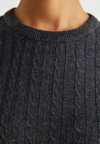 Gros plan d'une personne portant un pull en maille torsadée gris foncé avec un col rond côtelé.