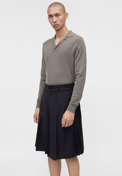 Άνδρας που φοράει γκρι μακρυμάνικο πλεκτό πόλο και μαύρη πλισέ φούστα μήκους μέχρι το γόνατο, σε απλό λευκό φόντο.