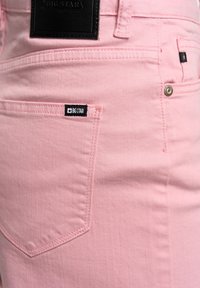 Gai rozā džinsi, izgatavoti no denima, ar melnu ādas piegriezpi un mazu melnu marķējumu netālu no kabatas. Redzami izteikti šuvumu detaļas.