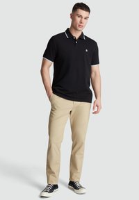 Zwarte polo met witte rand bij de kraag en mouwen, gecombineerd met beige broek en zwarte sneakers. Glad materiaal, klassieke pasvorm.