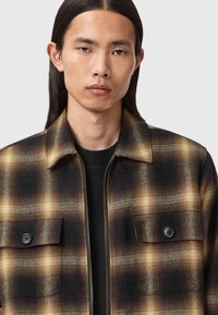 Camicia a quadri in tonalità di nero e marrone, design strutturato con tasche sul petto a bottone, chiusura con zip e un aspetto testurizzato simile alla lana.