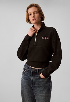 Ung kvinde iført en sort sweatshirt med halv lynlås og "Calvin Klein" logo og blå jeans, stående mod en ensfarvet grå baggrund.