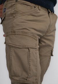 Khaki cargo kalhoty s několika kapsami, pevnými švy a tkanou strukturou. Zip na předním dílu s knoflíkovým zapínáním.