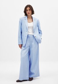 Ljusblå kostymjacka med spetsiga slag, dubbla knappar och vida matchande byxor. Vit T-shirt under och mönstrade mörka skor.