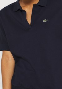 Marineblå pikétröja med V-ringning, korta ärmar och en texturerad tygkvalitet. Utrustad med en liten grön krokodil-logotyp på bröstet.