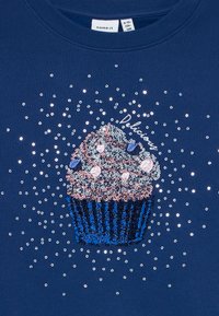 Zila džemperis ar kūciņas dizainu, ietverot spīdīgas sekundi, ar rozā krēmu un zilu iepakojumu, ko apņem mazāki sudrabaini sekundi.