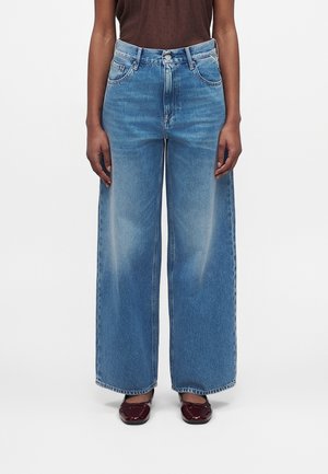 Personne portant un jean bleu taille haute à jambes larges, associé à des chaussures plates brillantes rouge foncé et un haut marron translucide à pois.