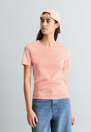 Młoda kobieta w brzoskwiniowej koszulce, odwróconej czapce z daszkiem, srebrnym łańcuszku i niebieskich dżinsach, stojąca na tle jednolitego jasnoszarego tła.