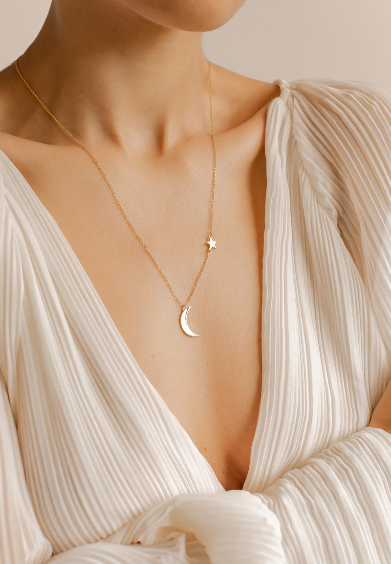 Collier en or arborant un pendentif en forme de croissant de lune et d'étoile, suspendu à une chaîne délicate, présenté sur un vêtement texturé blanc.