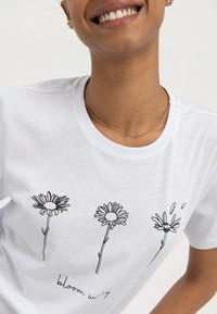 Bílý bavlněný tričko s černými konturami tří květů sedmikrásky a ručně psaným textem "bloom wi" pod nimi.