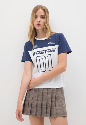 T-shirt a maniche corte con blocchi di colore blu navy e bianco, "BOSTON 01" stampato in blu navy. Abbinato a una gonna a pieghe a quadri marrone.